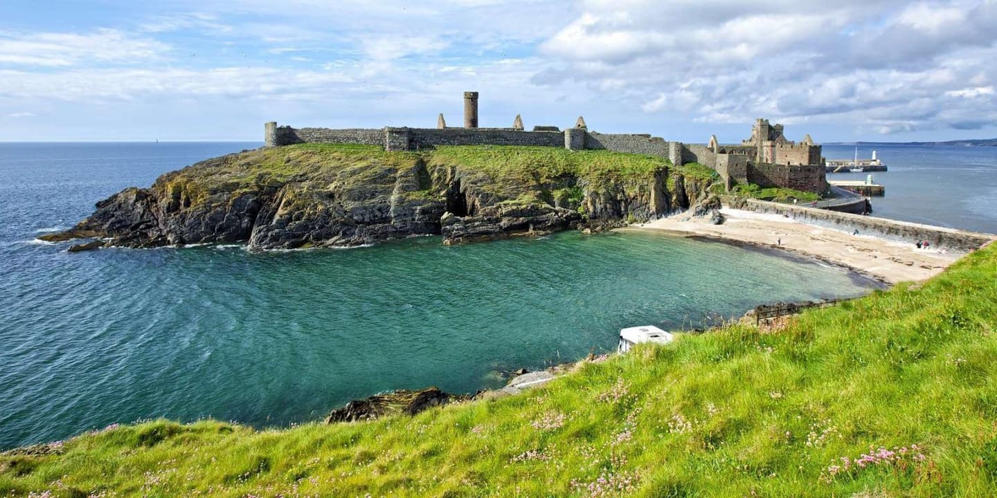 Peel Castle on the Isle of Man, situated on a rocky headland overlooking the sea. The ancient stone walls and tower stand above the green grass, with the ocean and a beach in the background.