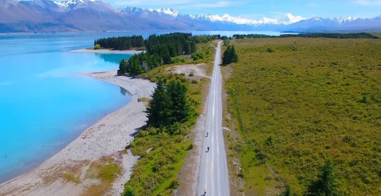 Cyclists riding on a long road between the ocean and a forest, with mountains in the distance