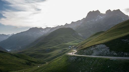 A cyclist rides along a winding road through the lush green valleys of Col de la Croix de Fer in the French Alps, with rugged mountains in the background under a dramatic sky.