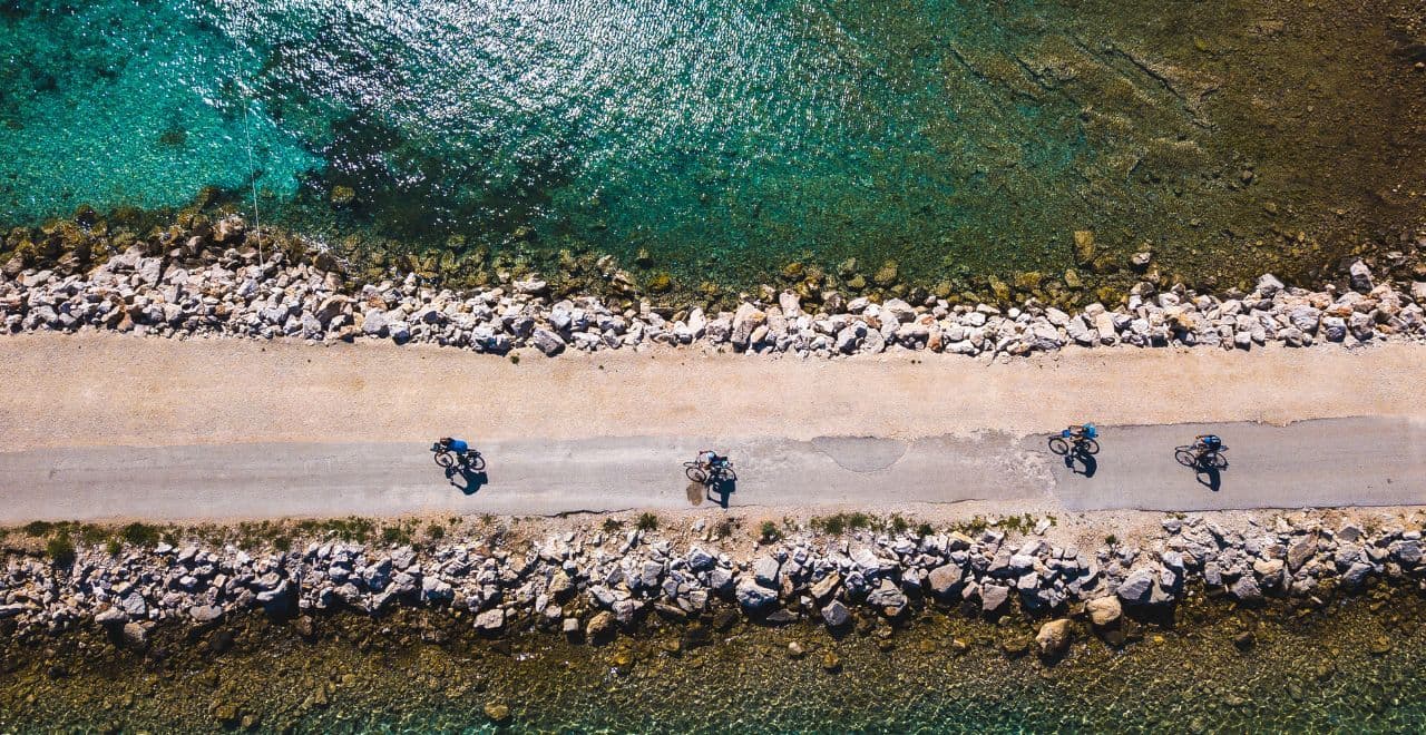 Aerial view of cyclists on a narrow coastal road, flanked by rocky shores and turquoise waters, ideal for adventurous cyclists
