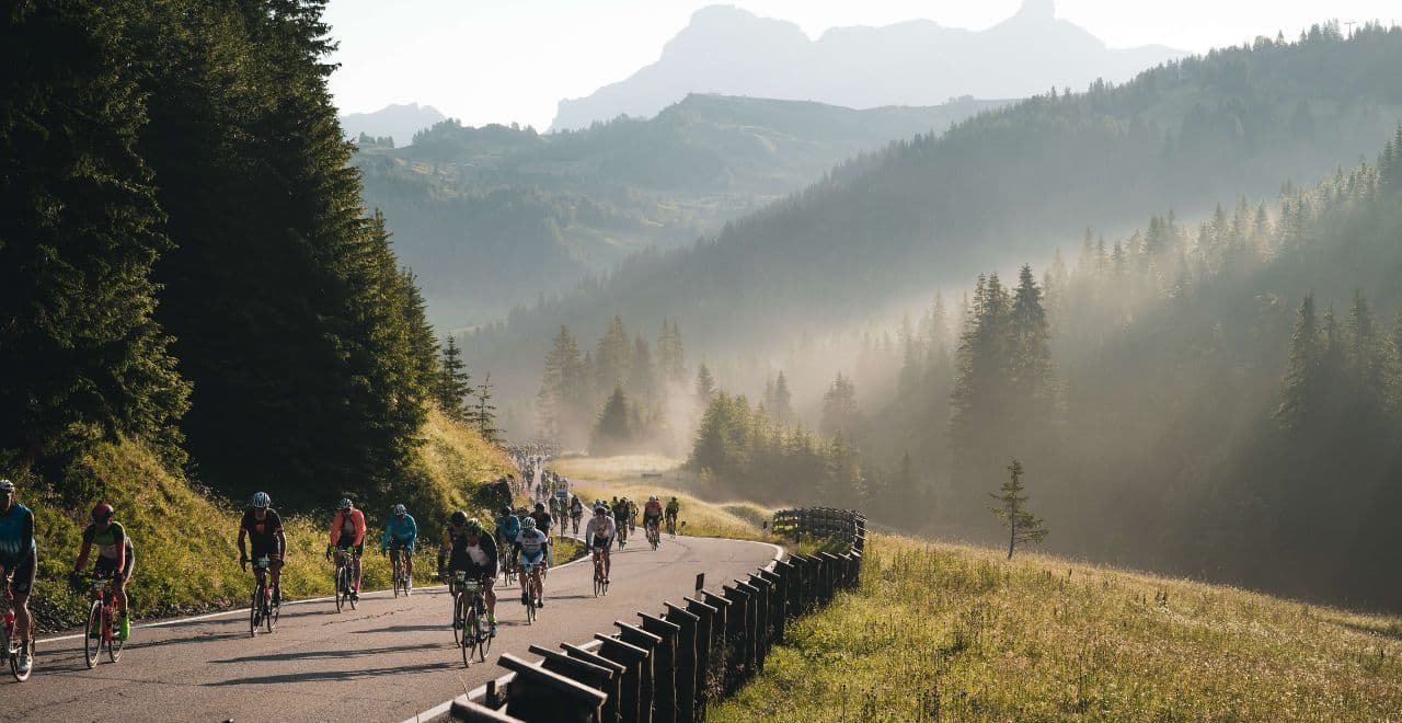 Cyclists riding on a winding mountain road in the Dolomites, surrounded by dense forests and mountain peaks in the distance.