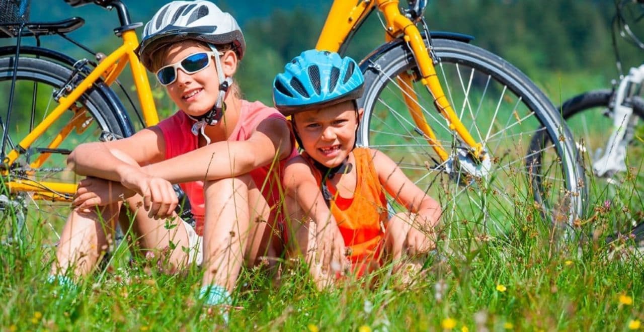 Two kids in helmets sitting beside bikes in grassy field