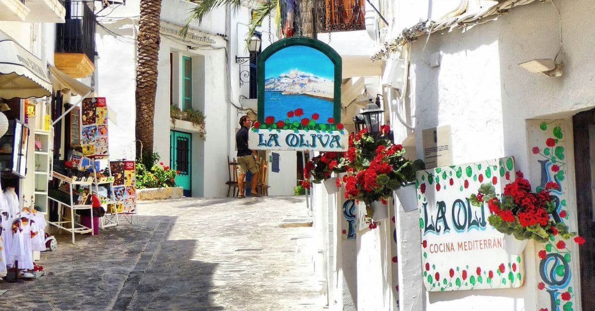 Charming white-washed alley with colorful pottery and a sign for 'La Oliva' restaurant in a sunny Mediterranean village