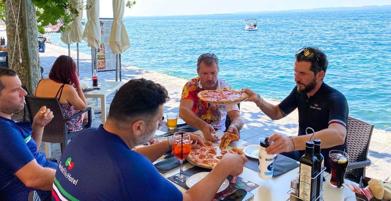 Group of cyclists sitting at a table by the water, sharing pizzas and drinks