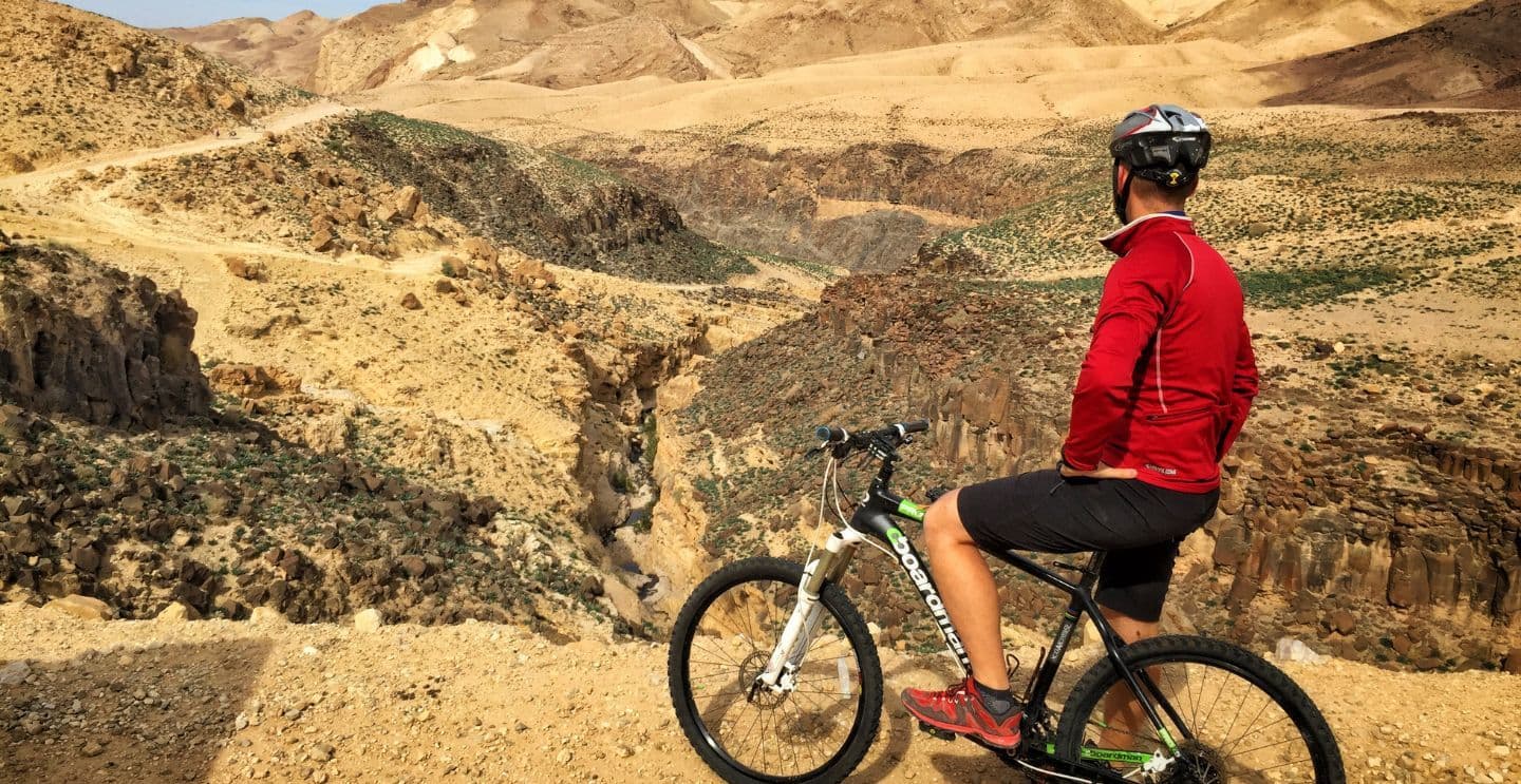 Cyclist in a red jacket pausing to admire a rugged canyon landscape with hills and rocky