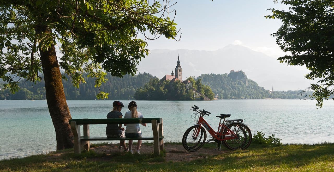 Cyclists resting on bench with view of Bled Island church across the lake
