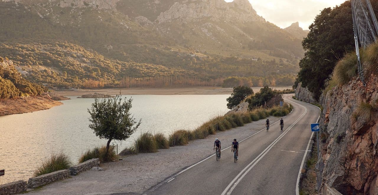 4 cyclists riding 2 abreast along a flat road next to a reservoir in Mallorca with mountains in the background