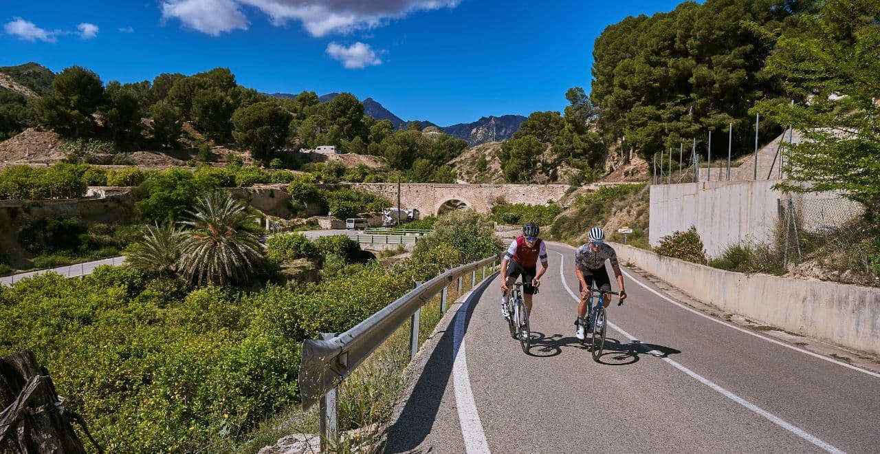 wo cyclists ride on a quiet road flanked by lush greenery and quaint houses under a bright blue sky.
