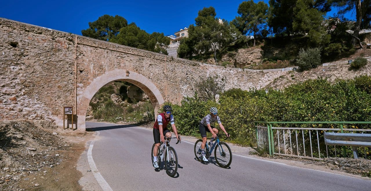 Two cyclists pass under an ancient stone arch bridge on a sunny day, with greenery and trees in the background.