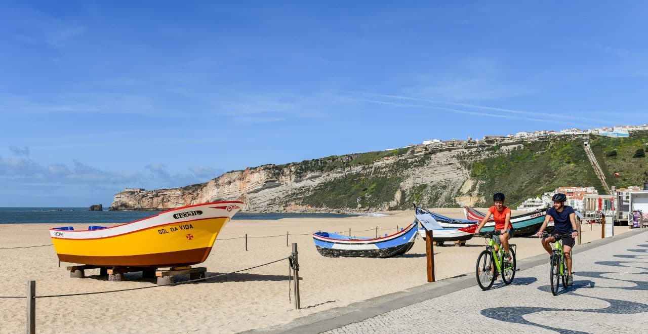 2 cyclists riding along a beach promenade with yellow boat on the sandy beach in front of them