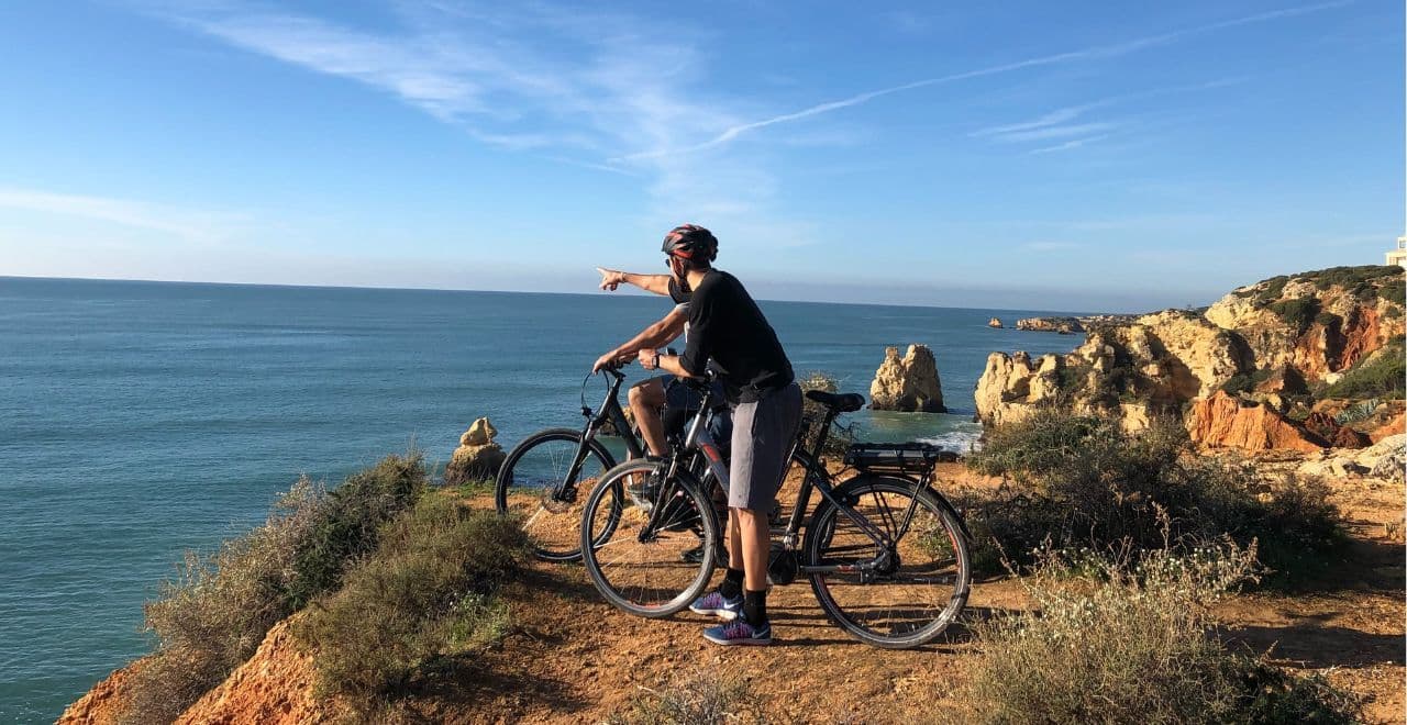 Two cyclists on a cliffside trail, one pointing out towards the horizon over the ocean.