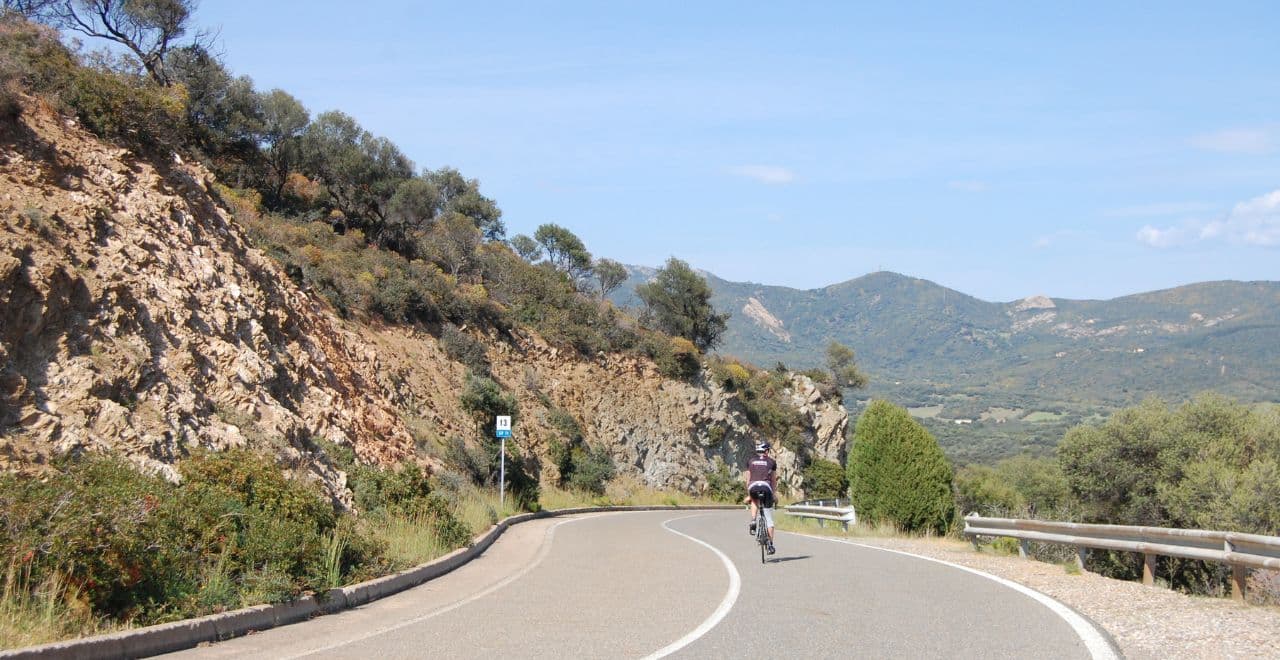 Cyclist riding on a mountain road with rocky hills and greenery.