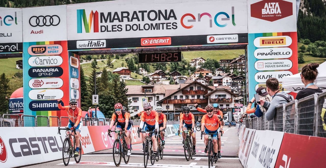 Cyclists crossing the finish line at the Maratona dles Dolomites event, with a backdrop of mountains and cheering spectators