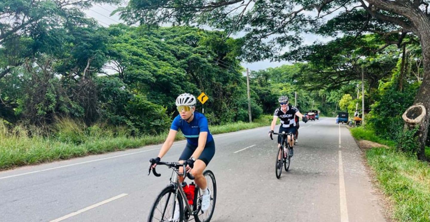 Two cyclists riding on a paved road surrounded by lush greenery and trees.