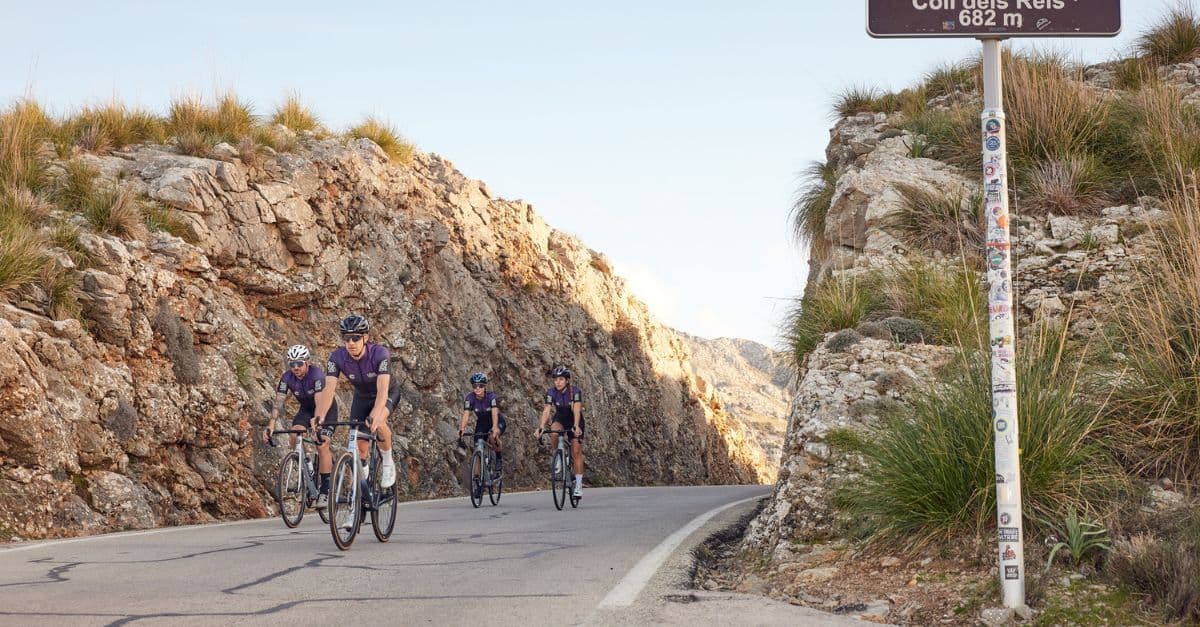 Cyclists pedaling up a steep incline, passing a signpost that marks their high-altitude achievement