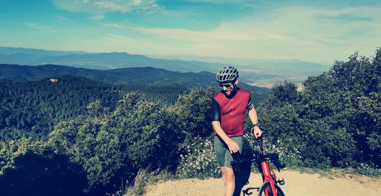 male cyclist standing with bike at top of a hill with lush forest and mountains in the background