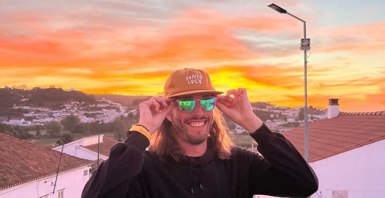 Smiling man in cap and sunglasses standing before vibrant sunset over hillside village.