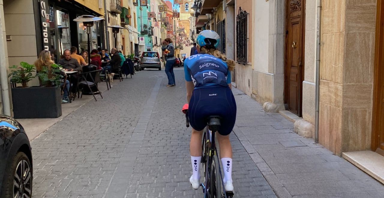 Cyclist riding through a narrow cobblestone street lined with cafes and shops.