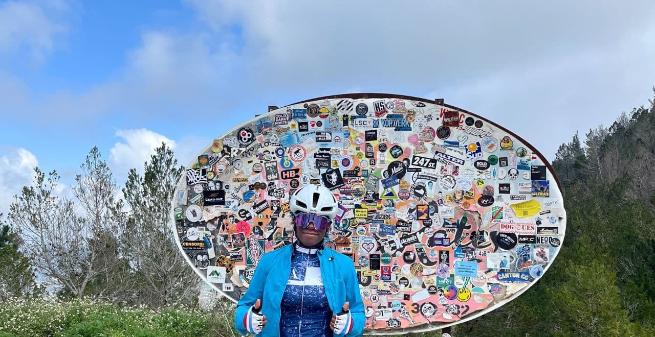 Cyclist in blue jacket posing in front of a large sticker-covered signboard on a mountain trail