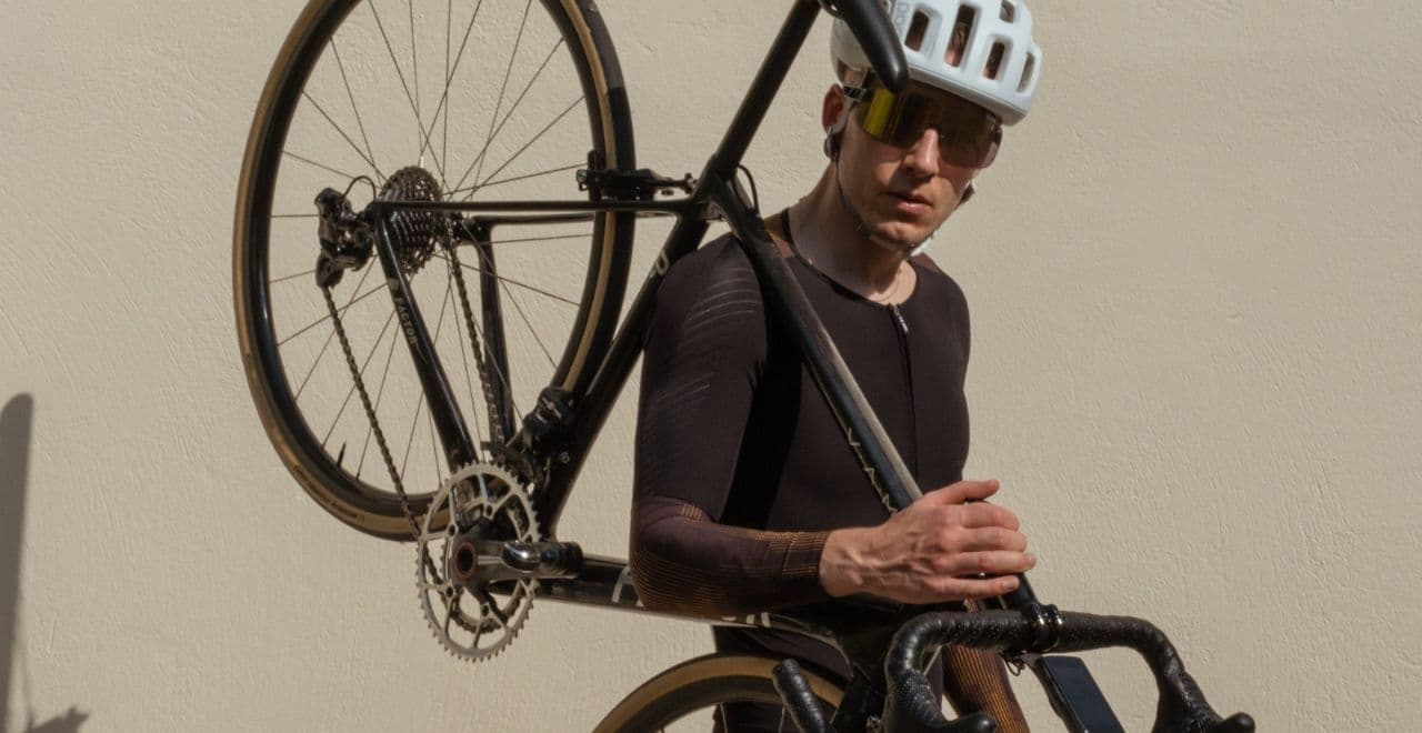 Cyclist in black kit carrying black road bike against beige wall background.