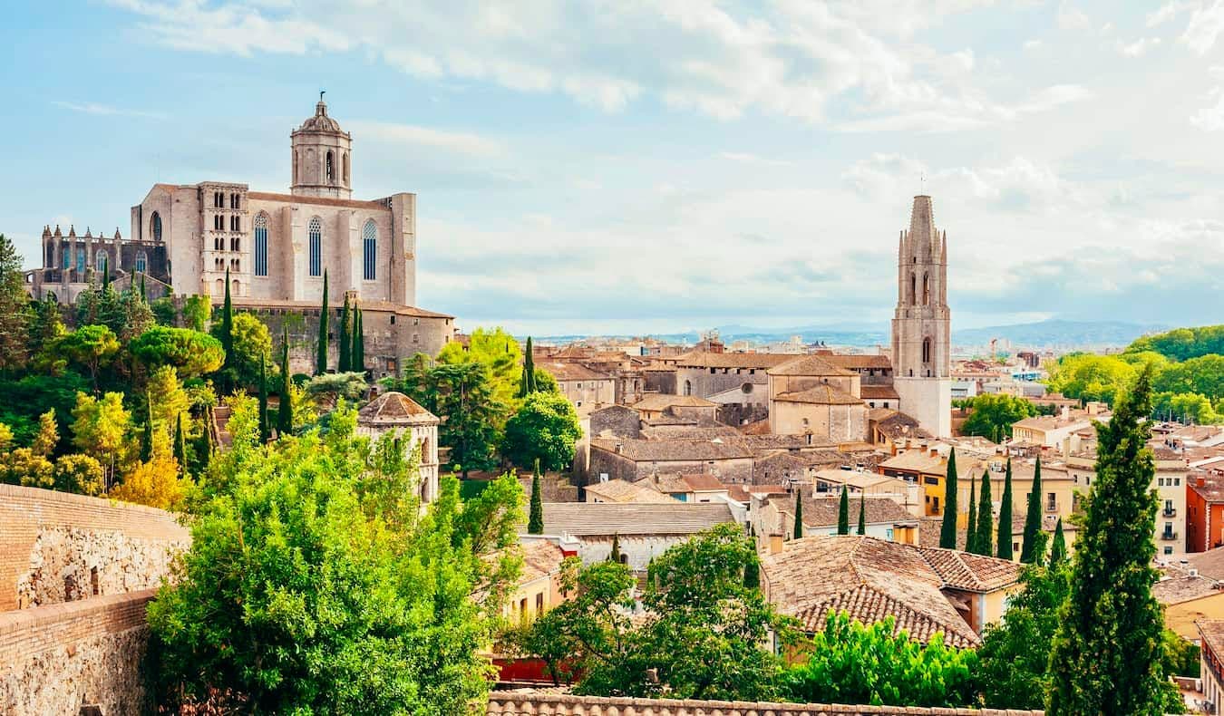 A panoramic view of Girona's old town, featuring the Girona Cathedral and the Basilica of Sant Feliu, surrounded by lush greenery and historic buildings.