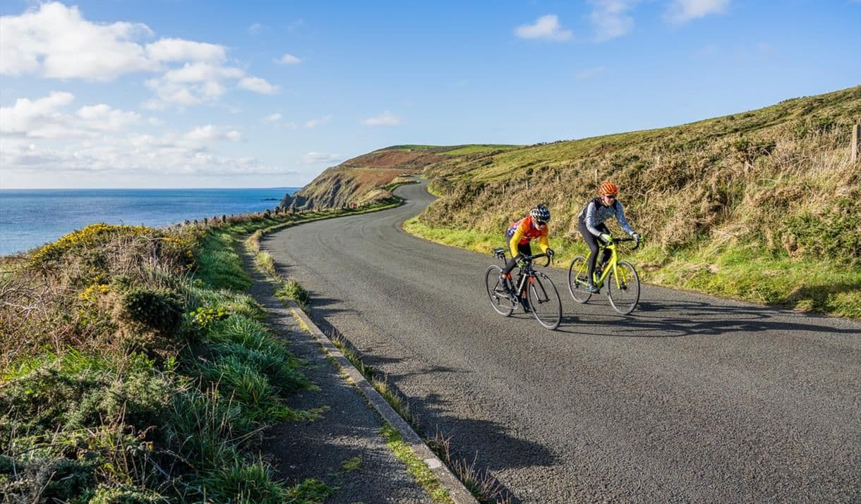 Two cyclists in vibrant gear riding on a sunny day along a scenic coastal road in the Isle of Man, with the ocean and cliffs in the background.