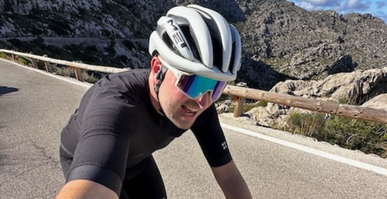 Cyclist wearing a white helmet and reflective sunglasses riding uphill on a mountain road.