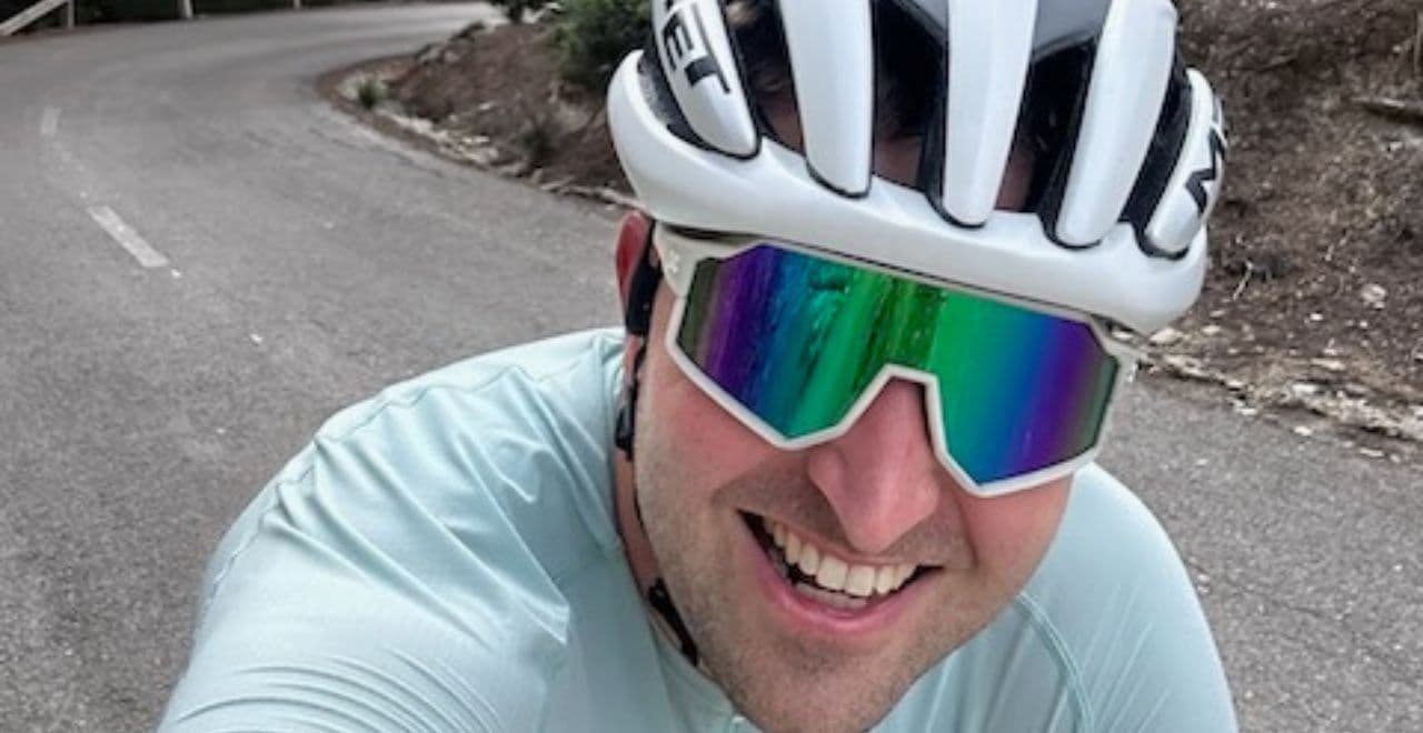 Smiling cyclist in a white helmet and mirrored sunglasses riding on a curving mountain road.