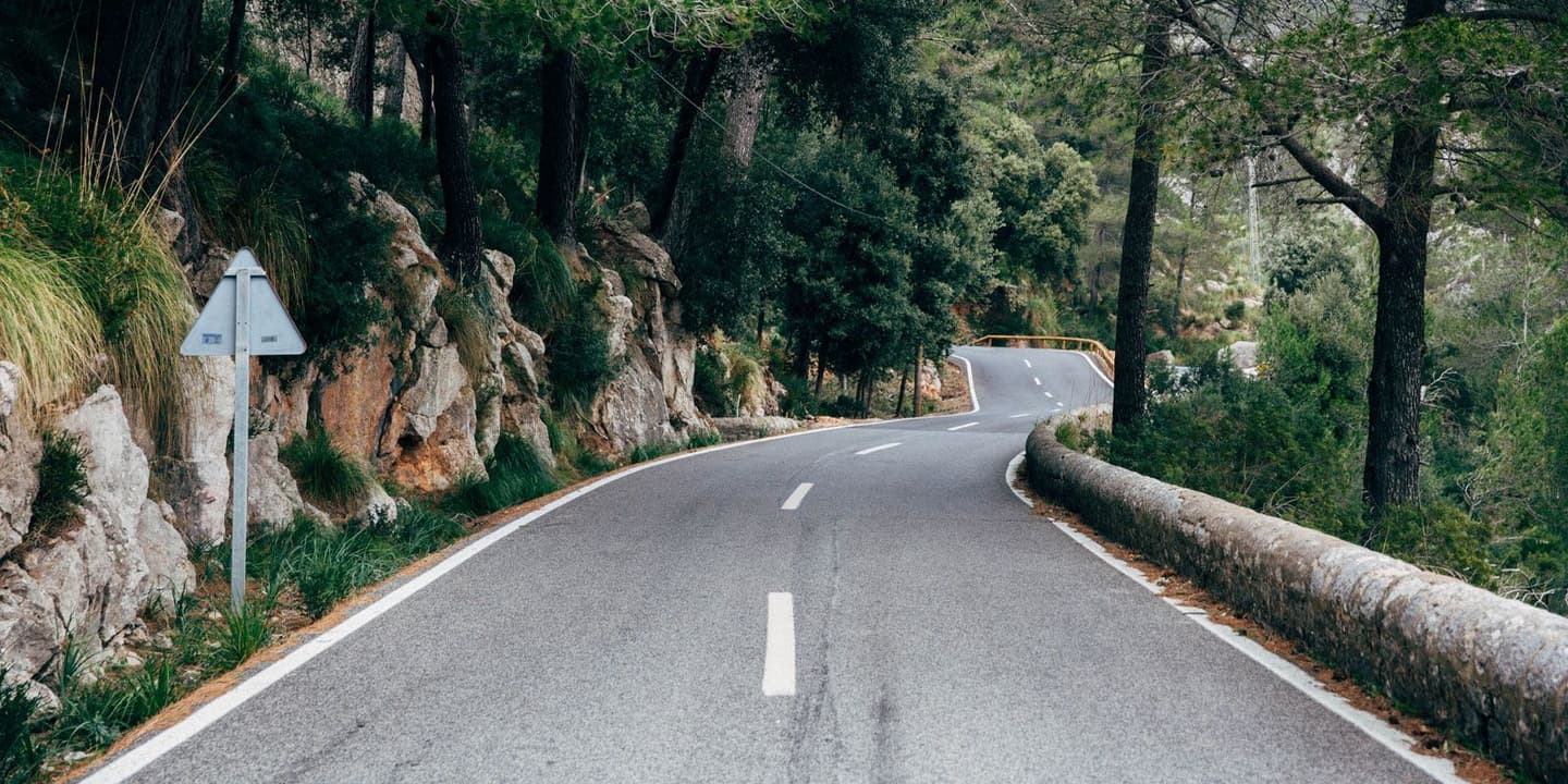 Winding mountain road through a forested area with rock formations and lush greenery in the Coll de sa Batalla, Mallorca.
