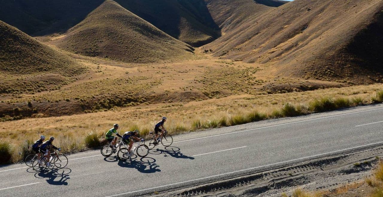 5 road cyclists riding on a road with mountains and hills to their side