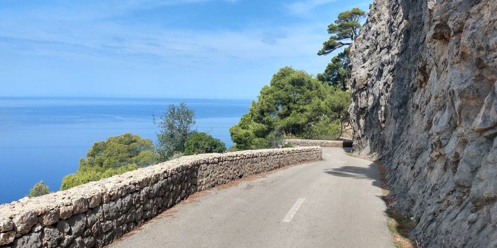 Narrow mountain road with stone walls winding along a cliffside, offering a scenic view of the Mediterranean Sea and lush green trees under a clear blue sky.